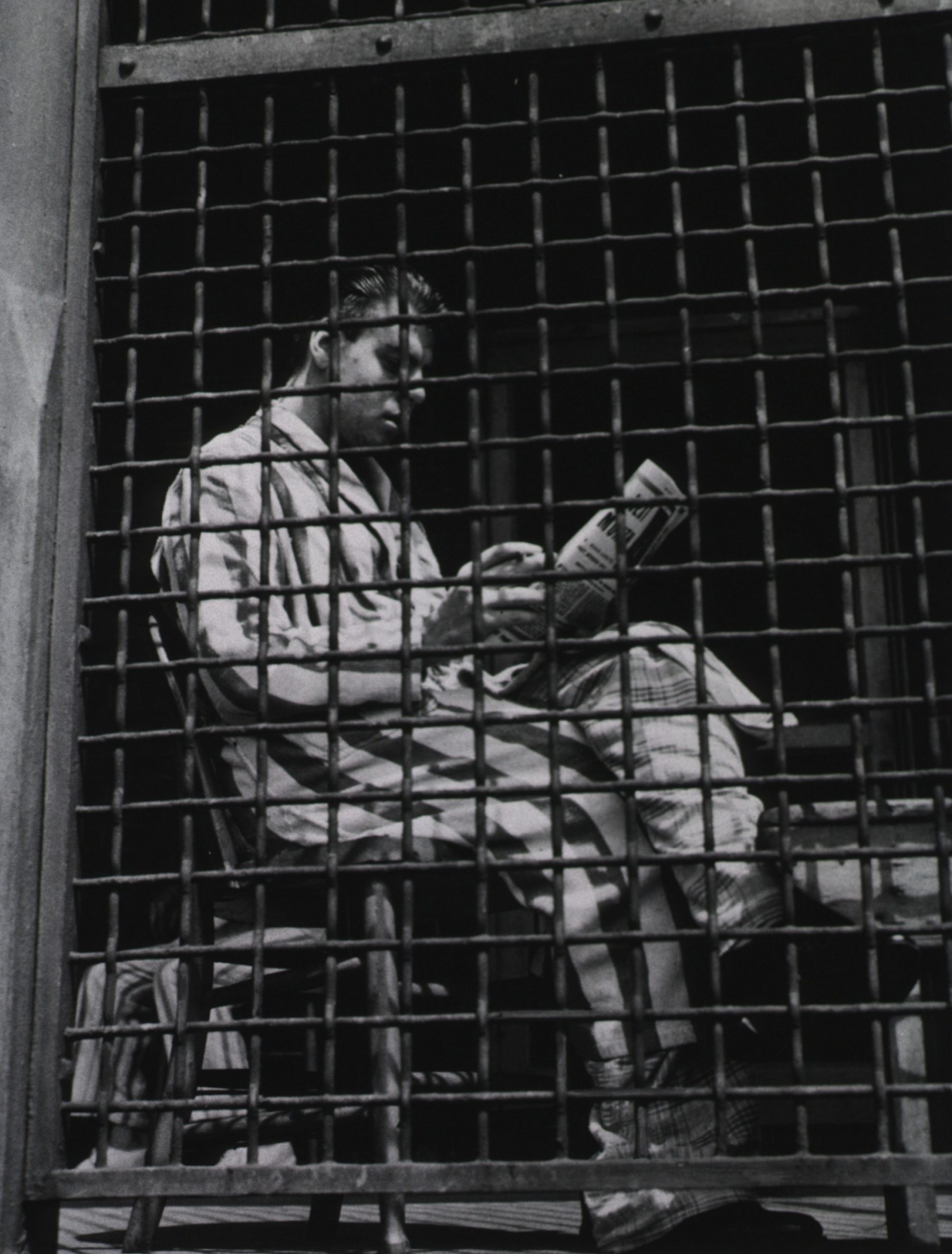 A man in pajamas and a robe is sitting and reading a newspaper behind a caged door in quarantine detention. Ellis Island, c 1930 h/t: NLM